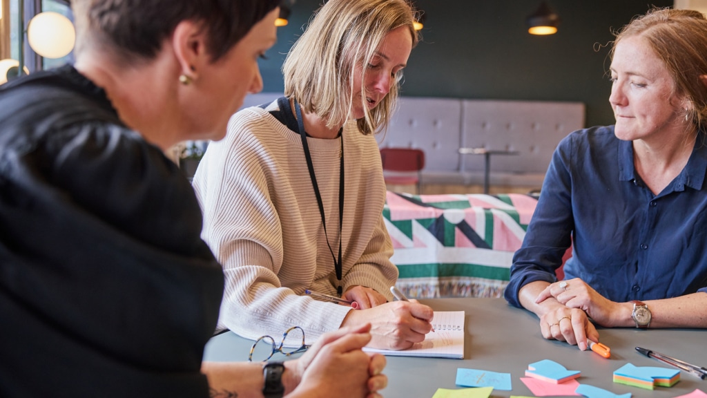 three women look to be in discussion around a table while one, a blonde woman with a bob, writes in a notebook