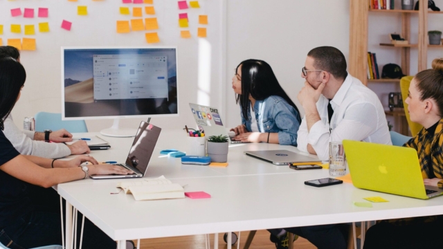 five people sat at a desk with laptops all looking at one screen away from the camera