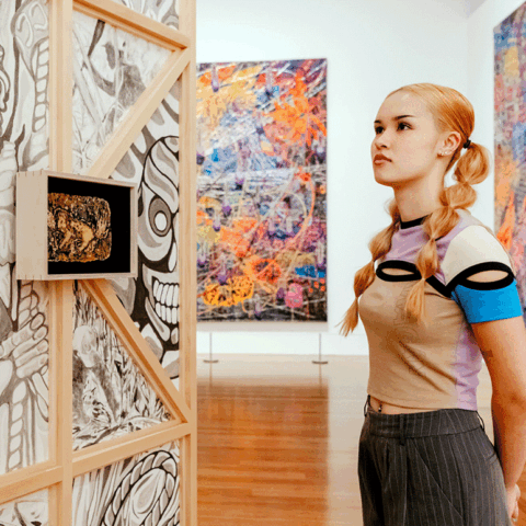 A young woman dressed in streetwear, with bubble braids in her blonde hair looks at a piece of artwork in a gallery, with large graphic canvases behind her