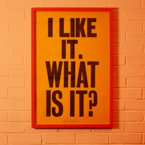 an orange poster on an orange wall with bold black text featuring the words I LIKE IT. WHAT IS IT?
