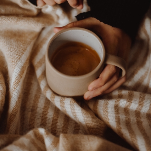 a mug of coffee being held by someone sat under a brown stripe blanket