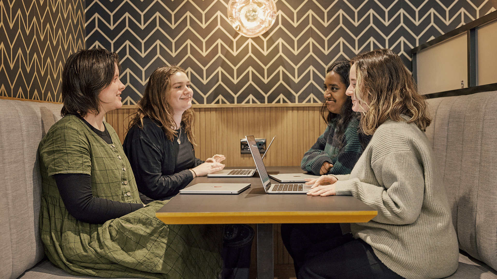 four women smile while sat at a table with laptops