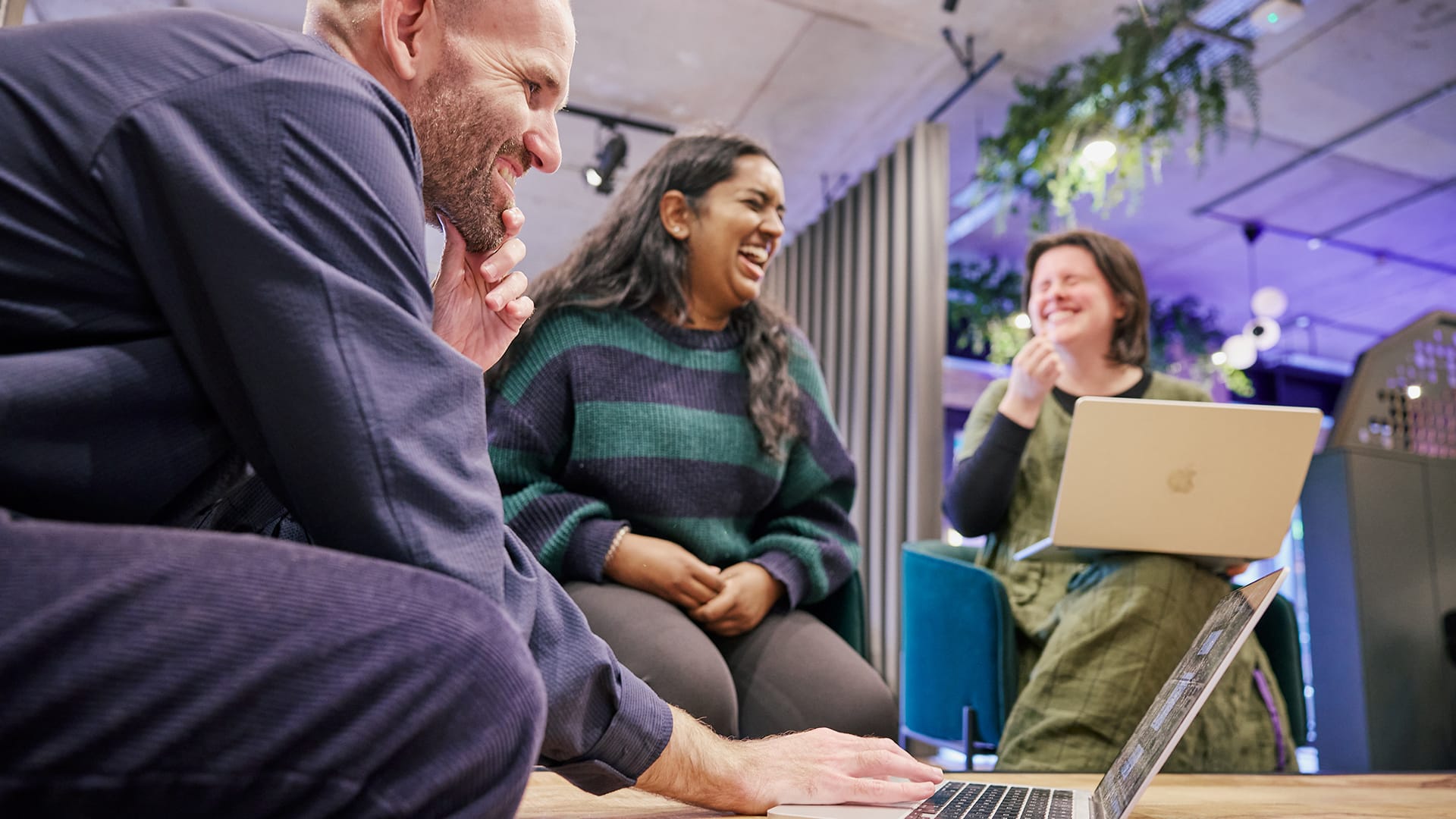 three people smiling, one man wearing a smart shirt and pants types on a laptop, a woman with tanned skin and black hair laughs while another woman with short brown hair wearing a green dress laughs along