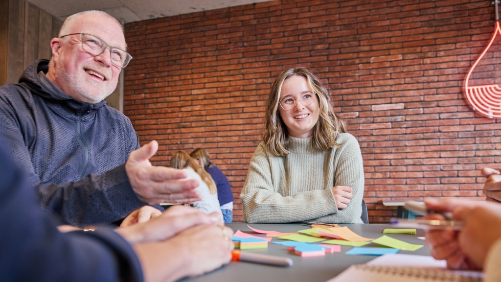 two people smile at the camera, one is an older balding man with glasses and a younger woman with short brown and blonde hair wearing a light green jumper sit at a table full of coloured post-it notes