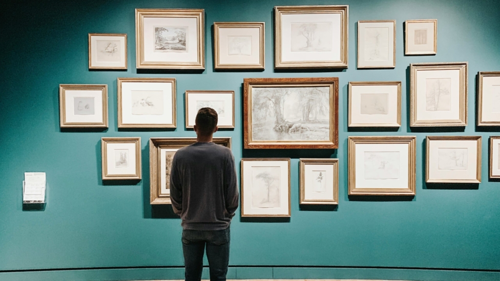 a man wearing jeans and a grey jumper looks away from the camera at a wall full of frames with sketches and line drawings in them