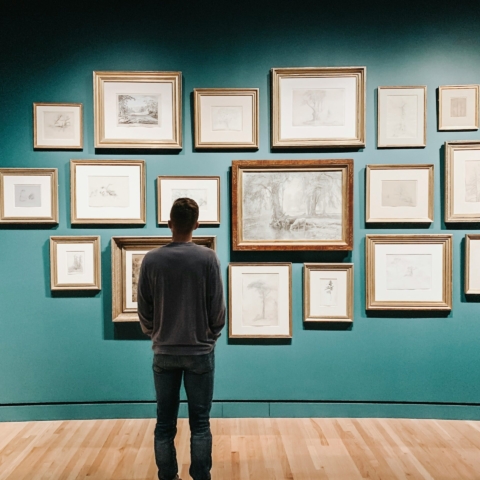 a man wearing jeans and a grey jumper looks away from the camera at a wall full of frames with sketches and line drawings in them