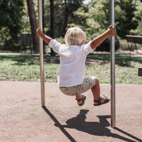 a small blonde child facing away from the camera, wearing a white shirt, grey shorts and sandals, holds onto two metal poles in a playground while jumping