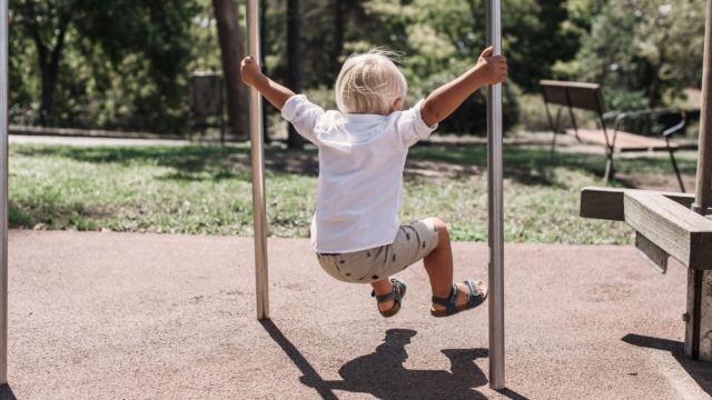 a small blonde child facing away from the camera, wearing a white shirt, grey shorts and sandals, holds onto two metal poles in a playground while jumping