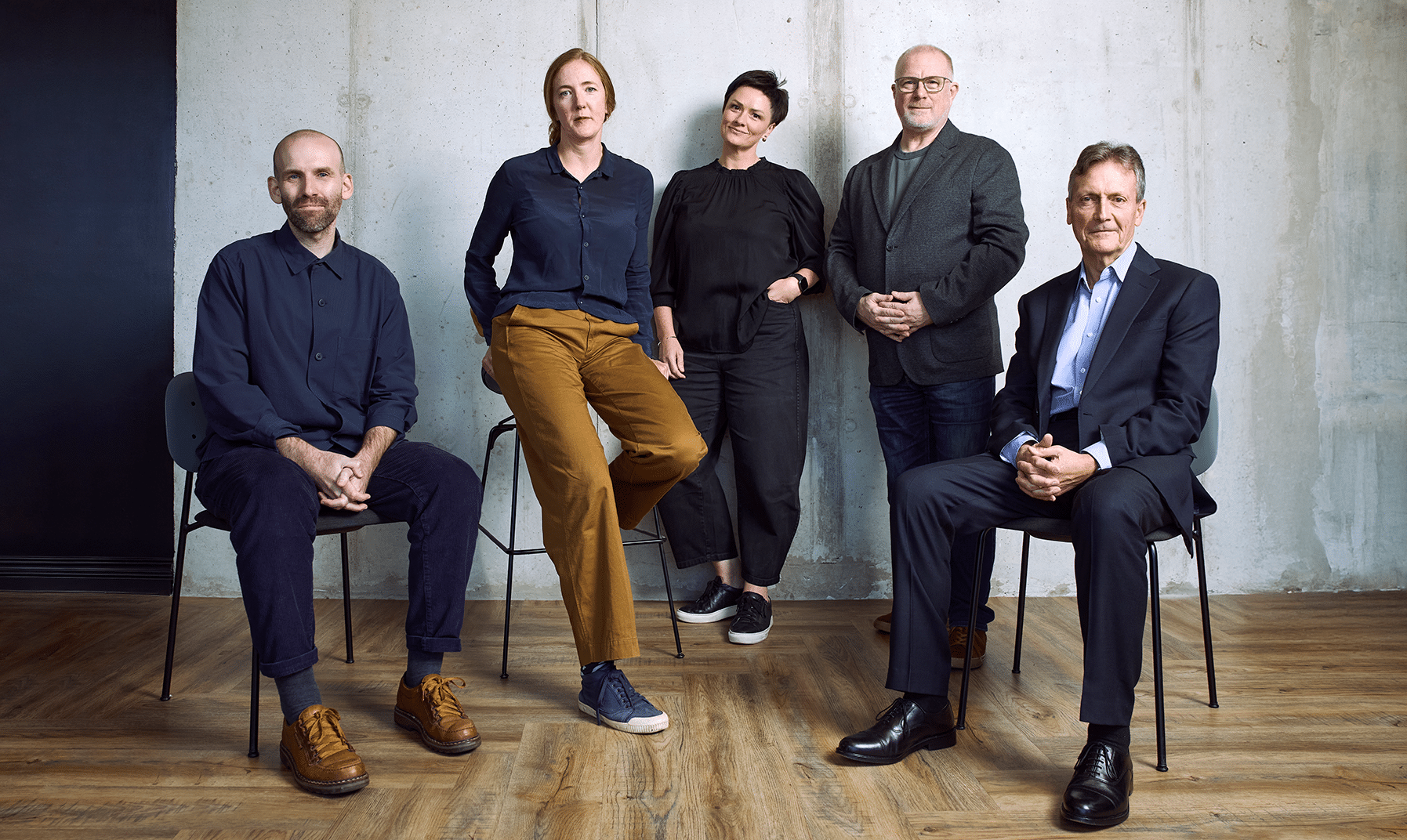 two women and three men all smartly dressed, some standing some sat on chairs all look at the camera. From left to right there is a bald man with stubble wearing a blue shirt, blue pants and light brown shoes, a red haired woman wearing a blue button up shirt with light brown pants and blue trainers, a woman with short brown hair wearing an all black outfit, a taller bald man with glasses and his hands crossed wears a charcoal grey suit jacket, and a man with dark grey hair wearing a full blue suit and polished black shoes