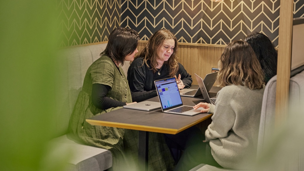 a group of four women sit around a small table, all with laptops, two are wearing black long sleeve t shirts