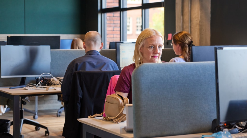a blonde woman with pink lipstick sits at a computer desk with a brown handhag on the desk